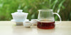 From left to right, a gaiwan, two teacups, and a glass server. The teacups and server have black tea in them.