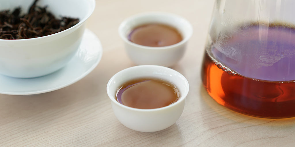 Two small teacups of black tea, with a gaiwan in the background on the left and a glass server on the right.