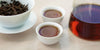 Two small teacups of black tea, with a gaiwan in the background on the left and a glass server on the right.