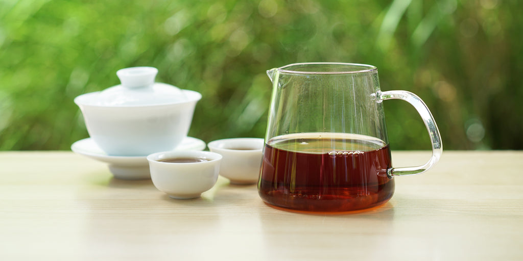 From left to right, a gaiwan, two teacups, and a glass server. The teacups and server have black tea in them.