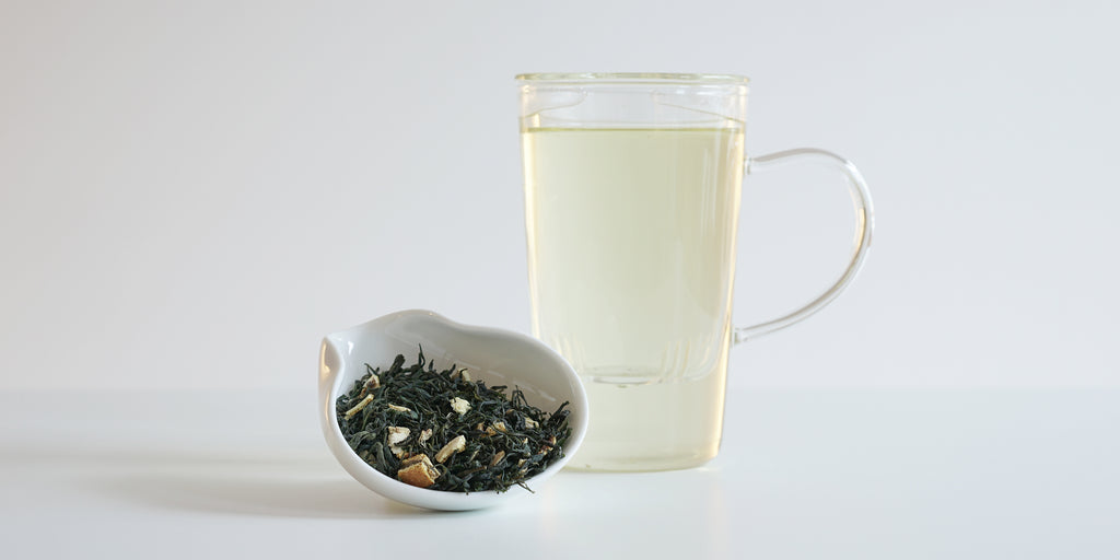 A white dish of dry green tea leaves, dried orange peel, and ginger. A glass cup of brewed tea is in the background.