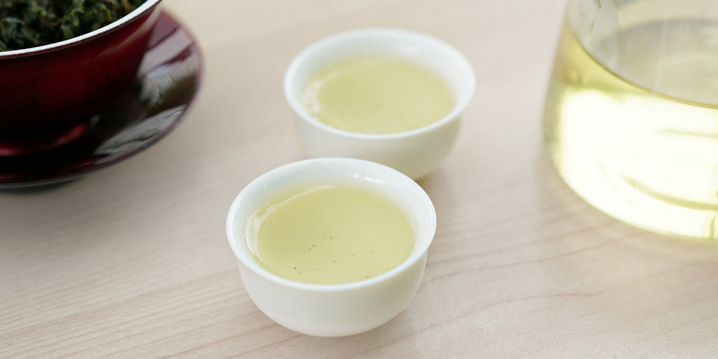 Two small teacups of Formosa oolong tea, with a gaiwan in the background on the left and a glass server on the right.