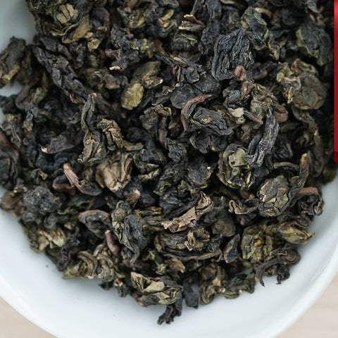 A white dish containing dry Tieguanyin tea leaves. A red ribbon in the top right corner reads “Red Label Collection”