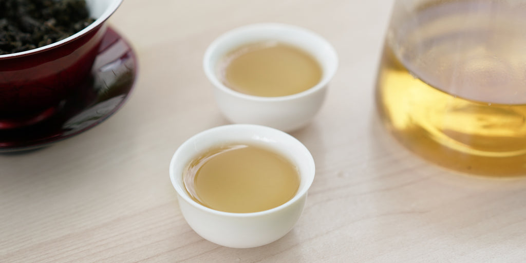 Two small teacups of Tieguanyin tea, with a gaiwan in the background on the left and a glass server on the right.