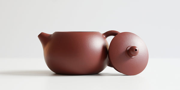 Side view of a terracotta-colored yixing teapot. The lid is facing the camera, resting against the handle.