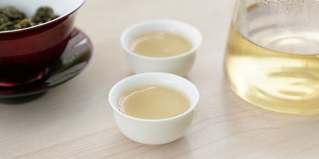 Two small teacups of jasmine tea, with a gaiwan in the background on the left and a glass server on the right.