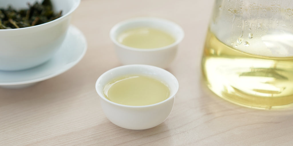 Two small teacups of Formosa oolong tea, with a gaiwan in the background on the left and a glass server on the right.