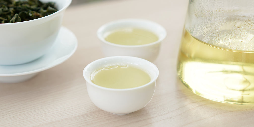 Two small teacups of Formosa oolong tea, with a gaiwan in the background on the left and a glass server on the right.