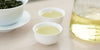 Two small teacups of Formosa oolong tea, with a gaiwan in the background on the left and a glass server on the right.