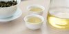 Two small teacups of Formosa oolong tea, with a gaiwan in the background on the left and a glass server on the right.