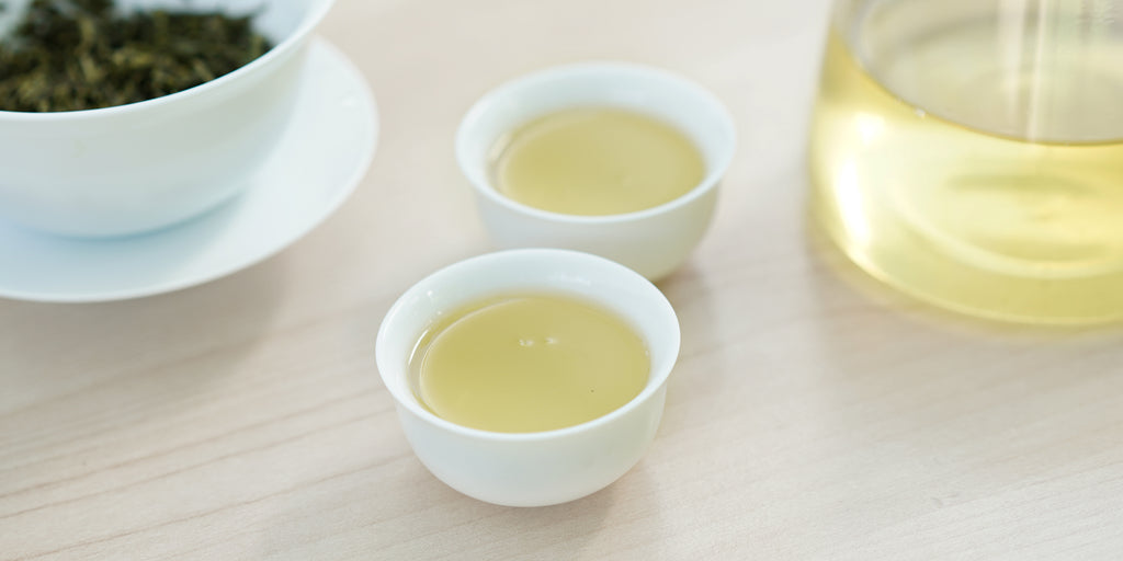 Two small teacups of green tea, with a gaiwan in the background on the left and a glass server on the right.