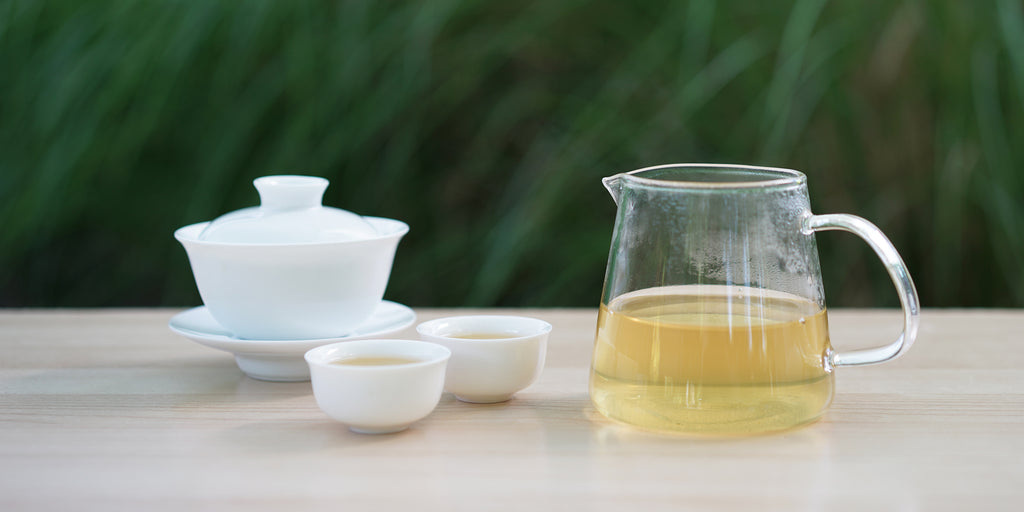 From left to right, a gaiwan, two teacups, and a glass server. The teacups and server have green tea in them.