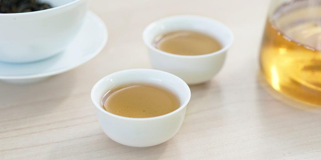 Two small teacups of green tea, with a gaiwan in the background on the left and a glass server on the right.
