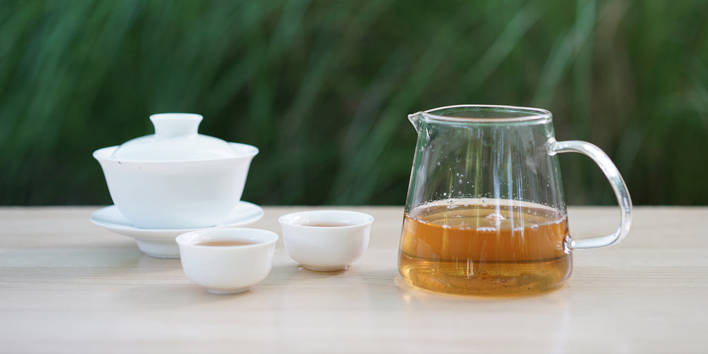 From left to right, a gaiwan, two teacups, and a glass server. The teacups and server have green tea in them.