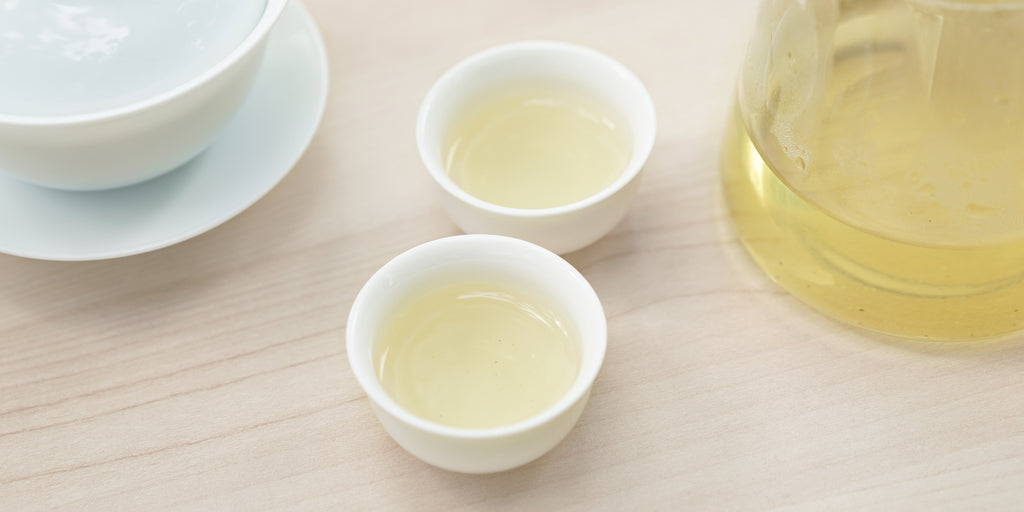 Two small teacups of green tea, with a gaiwan in the background on the left and a glass server on the right.