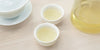 Two small teacups of green tea, with a gaiwan in the background on the left and a glass server on the right.