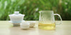 From left to right, a gaiwan, two teacups, and a glass server. The teacups and server have green tea in them.