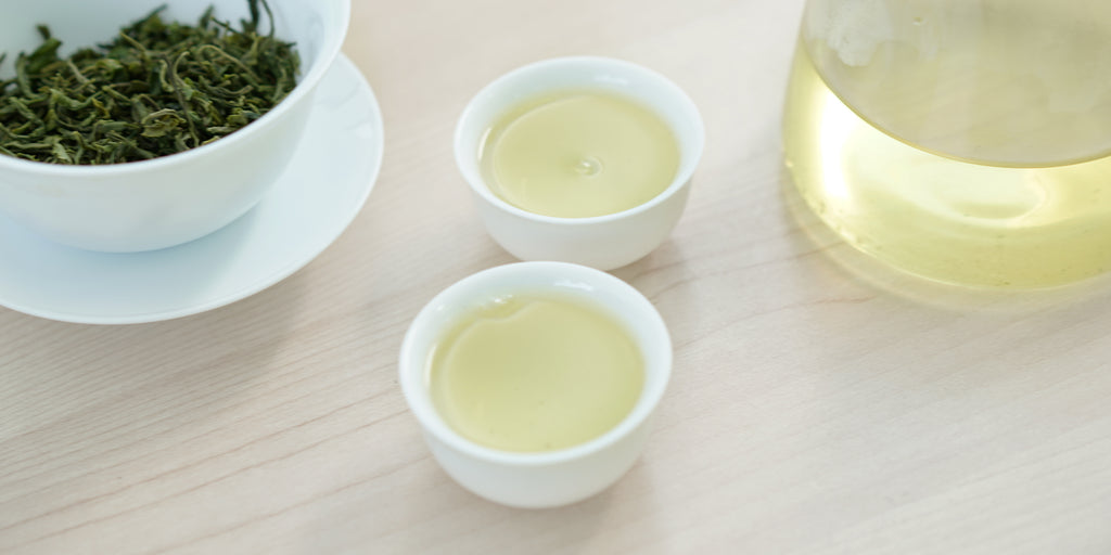 Two small teacups of green tea, with a gaiwan in the background on the left and a glass server on the right.