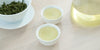 Two small teacups of green tea, with a gaiwan in the background on the left and a glass server on the right.
