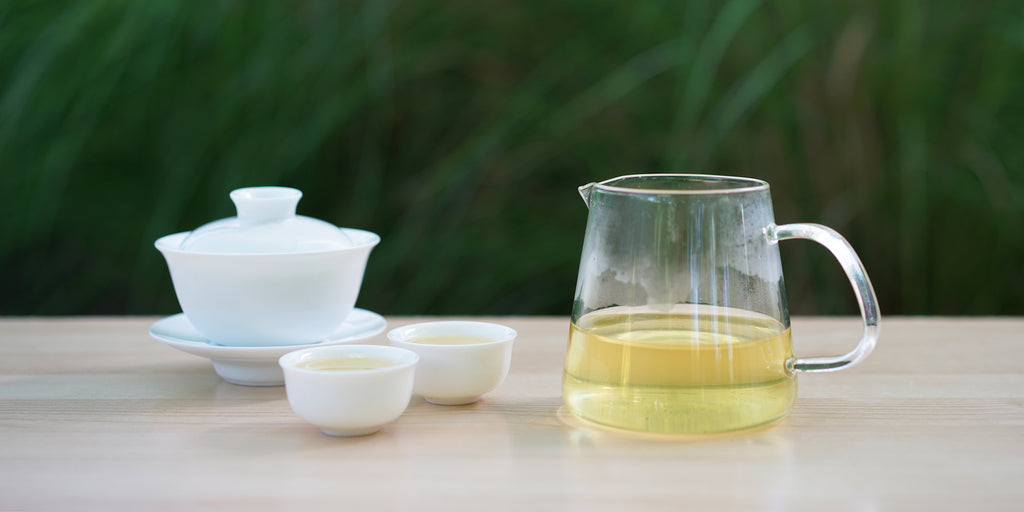 From left to right, a gaiwan, two teacups, and a glass server. The teacups and server have green tea in them.