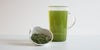 A white tea dish with dry green tea leaves, toasted rice and matcha powder. A glass cup full of brewed genmai matcha is in the background.