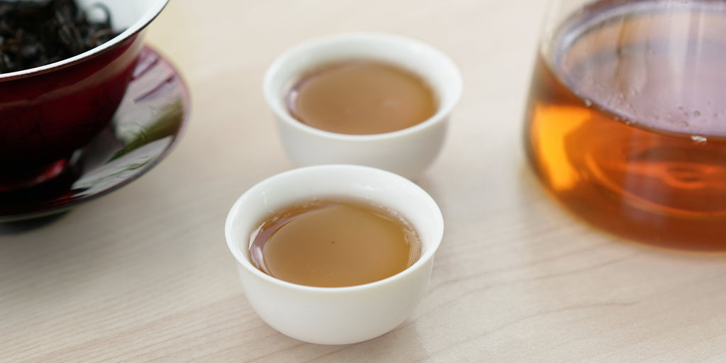 Two small teacups of Wuyi oolong tea, with a gaiwan in the background on the left and a glass server on the right.
