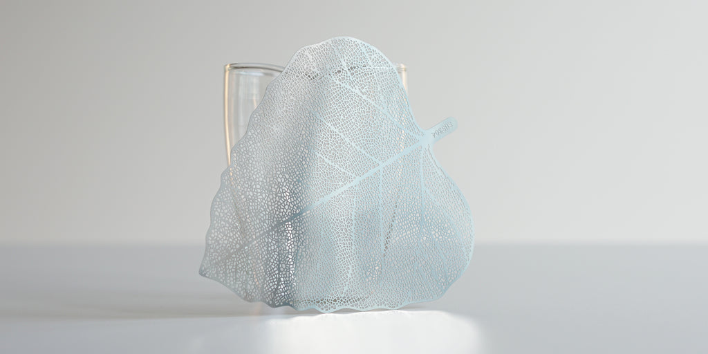 Steel leaf strainer in the shape of a bodhi leaf leaning against a glass server.