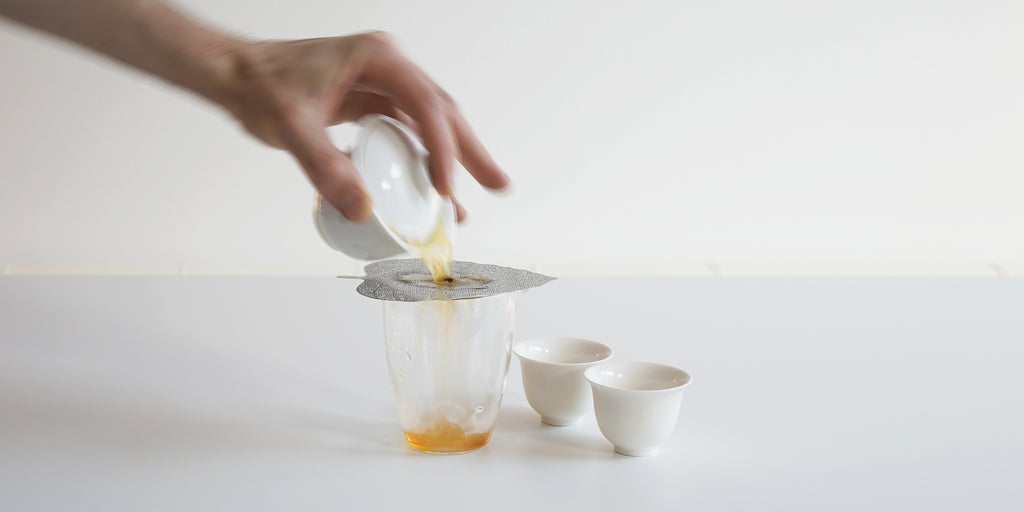 A hand in motion starting to pour a gaiwan, using a metal strainer placed over the glass server to catch any leaves.