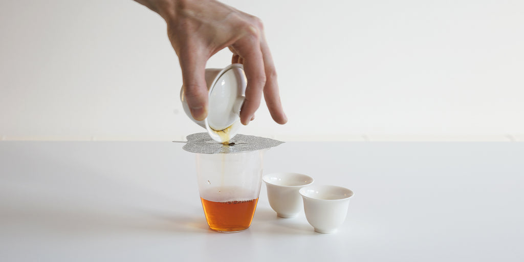 Metal strainer placed over a glass server being used to filter out leaves slipping through a gaiwan being poured from above.