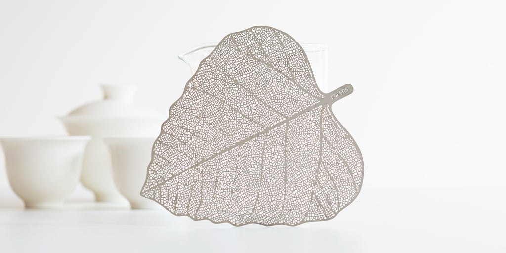 A steel strainer in the shape of a bodhi leaf leaned against a glass server. A pair of white cups and a gaiwan are in the background.