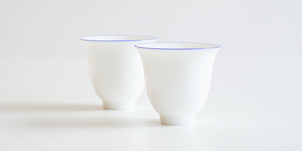 Side view of a pair of white porcelain teacups, with a ring of blue painted around the rim.