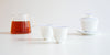 A pair of white teacups, with a glass server filled with tea and a gaiwan in the background.