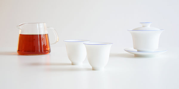 A pair of white teacups, with a glass server filled with tea and a gaiwan in the background.
