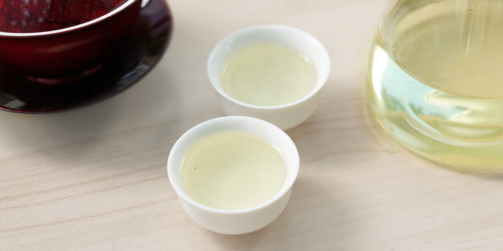 Two small teacups of green tea, with a gaiwan in the background on the left and a glass server on the right.