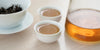 Two small teacups of Phoenix oolong tea, with a gaiwan in the background on the left and a glass server on the right.