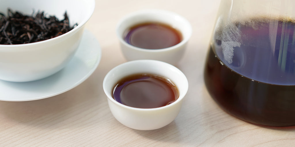 Two small teacups of pu-erh tea, with a gaiwan in the background on the left and a glass server on the right.