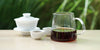 From left to right, a gaiwan, two teacups, and a glass server. The teacups and server have pu-erh tea in them.