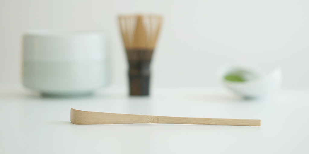 A matcha scoop set on a white surface, with a white bowl, matcha whisk, and a white dish full of matcha in the background.