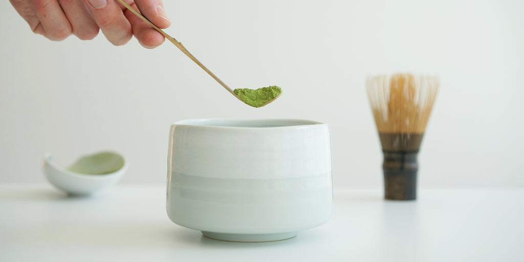 A hand holding a bamboo matcha scoop with matcha over a bowl. A white dish filled with matcha and a matcha whisk are in the background.