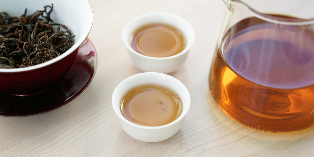Two small teacups of black tea, with a gaiwan in the background on the left and a glass server on the right.