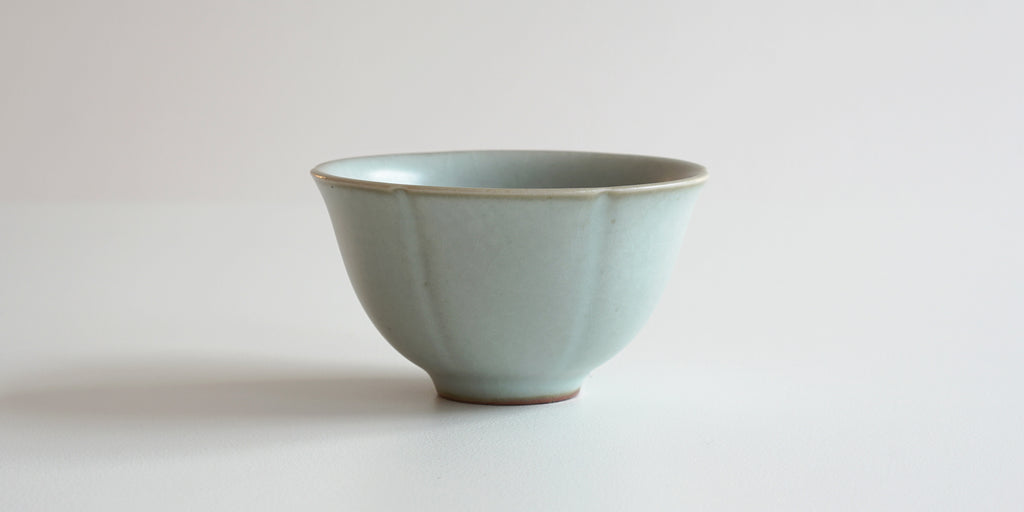 Side view of a blue-green celadon teacup. The outside has slight grooves in it to look like a lotus flower.