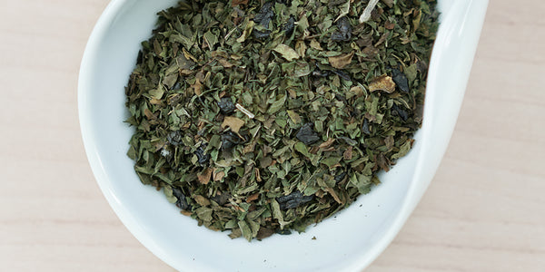 A white dish containing dry green tea and peppermint leaves.