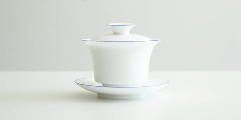 Side view of a white porcelain gaiwan, with blue rings painted around the edges of the plate, bowl, and the handle of the lid. The sides start straight before curving out towards the lip.