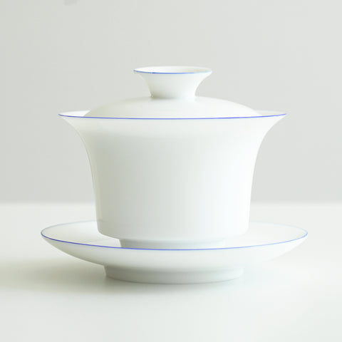 Side view of a white porcelain gaiwan, with blue rings painted around the edges of the plate, bowl, and the handle of the lid. The sides start straight before curving out towards the lip.