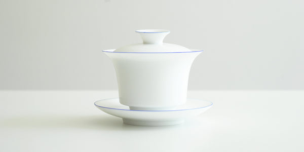 Side view of a white porcelain gaiwan, with blue rings painted around the edges of the plate, bowl, and the handle of the lid. The sides start straight before curving out towards the lip.