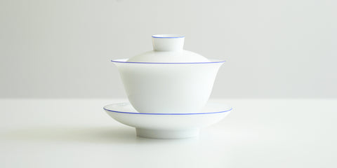 Side view of a white porcelain gaiwan, with blue rings painted around the edges of the plate, bowl, and the handle of the lid. The sides are rounded at the bottom before curving out towards the lip.