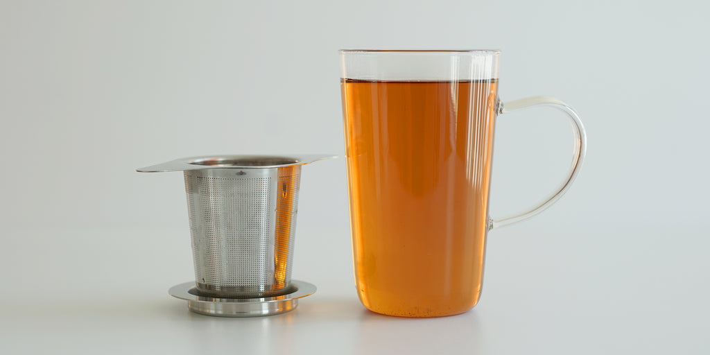 A steel infuser basket with two wings to hang on top of a cup rests on top of a steel plate. A glass cup full of tea stands next to it.