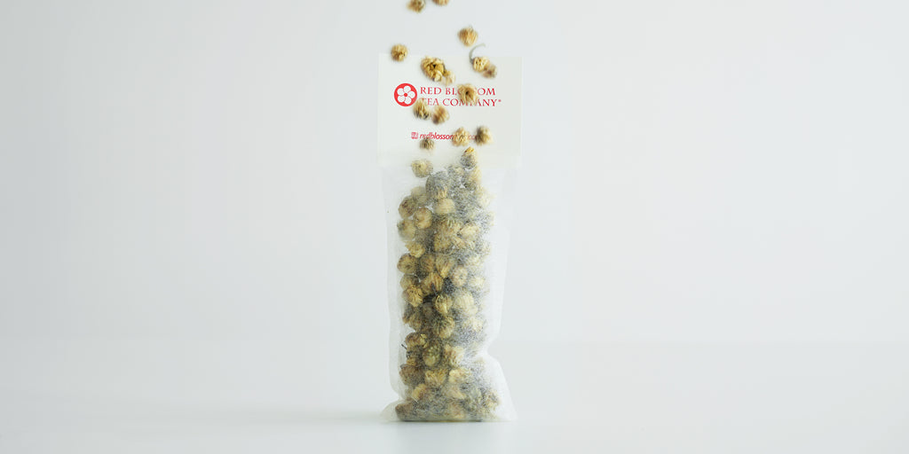 A tea sachet standing filled with chrysanthemum buds. More chrysanthemum buds are raining down from above.