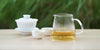 From left to right, a gaiwan, two teacups, and a glass server. The teacups and server have white tea in them.