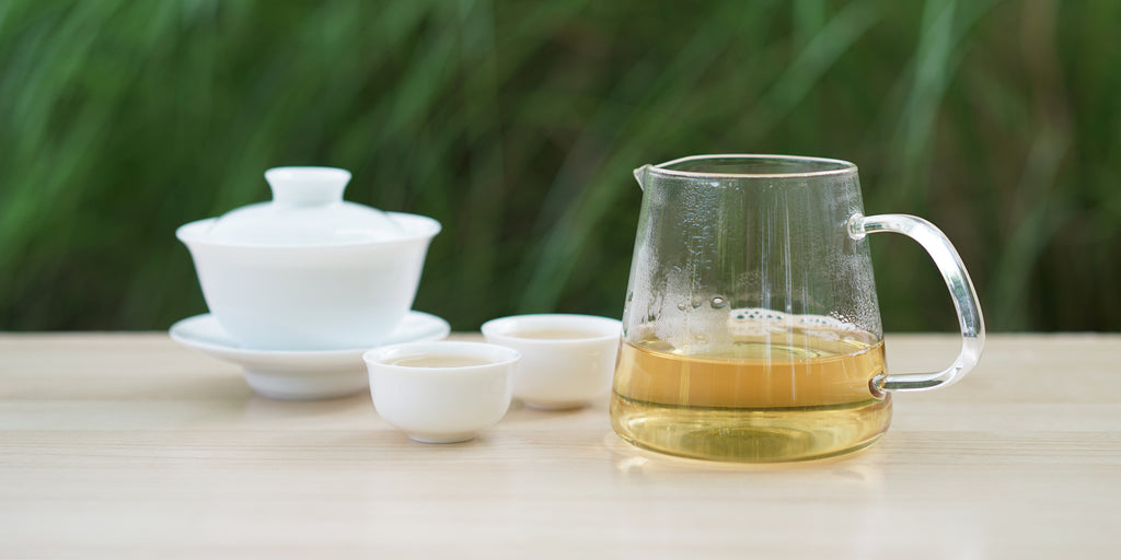 From left to right, a gaiwan, two teacups, and a glass server. The teacups and server have white tea in them.